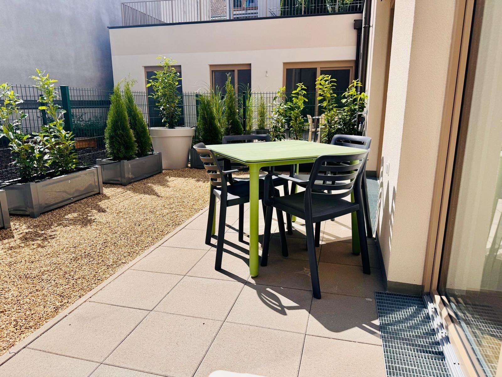 An outdoor patio with a table and chairs, surrounded by potted plants and a building in the background.