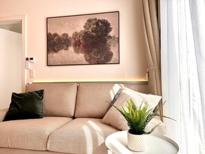 Ein modernes Wohnzimmer mit einem beige Colordesign, einem gerahmten Landschaftsbild, einer Topfpflanze und einem weißen Beistelltisch.