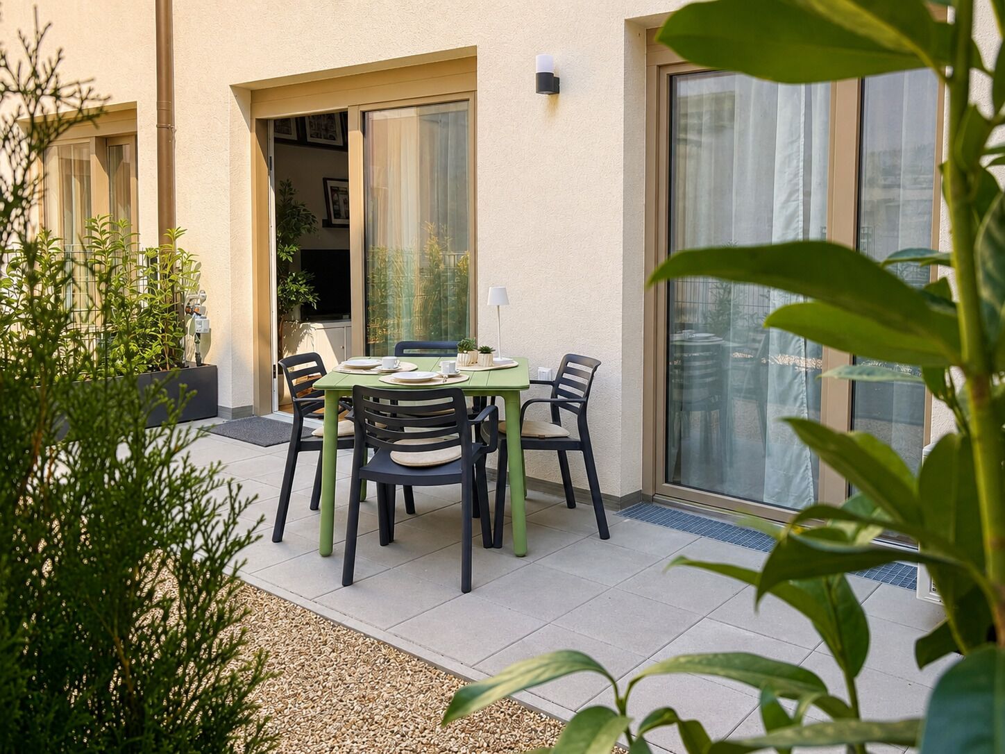 Une terrasse extérieure avec une table et des chaises, entourée de plantes et donnant sur l’entrée d’un bâtiment.