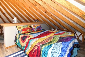 Ein gemütliches Schlafzimmer im Dachgeschoss mit Holzbalkendecke, einem Bett mit bunter Steppdecke und einem kleinen Tisch mit einer Tasse.