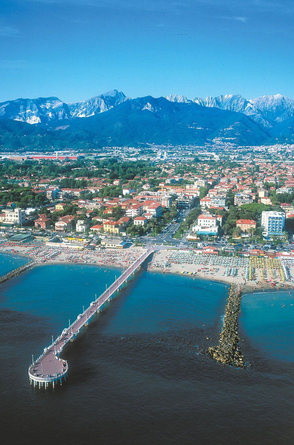 Eine Luftaufnahme einer Küstenstadt mit einer Mole, die sich ins Meer erstreckt, umgeben von Bergen im Hintergrund.
