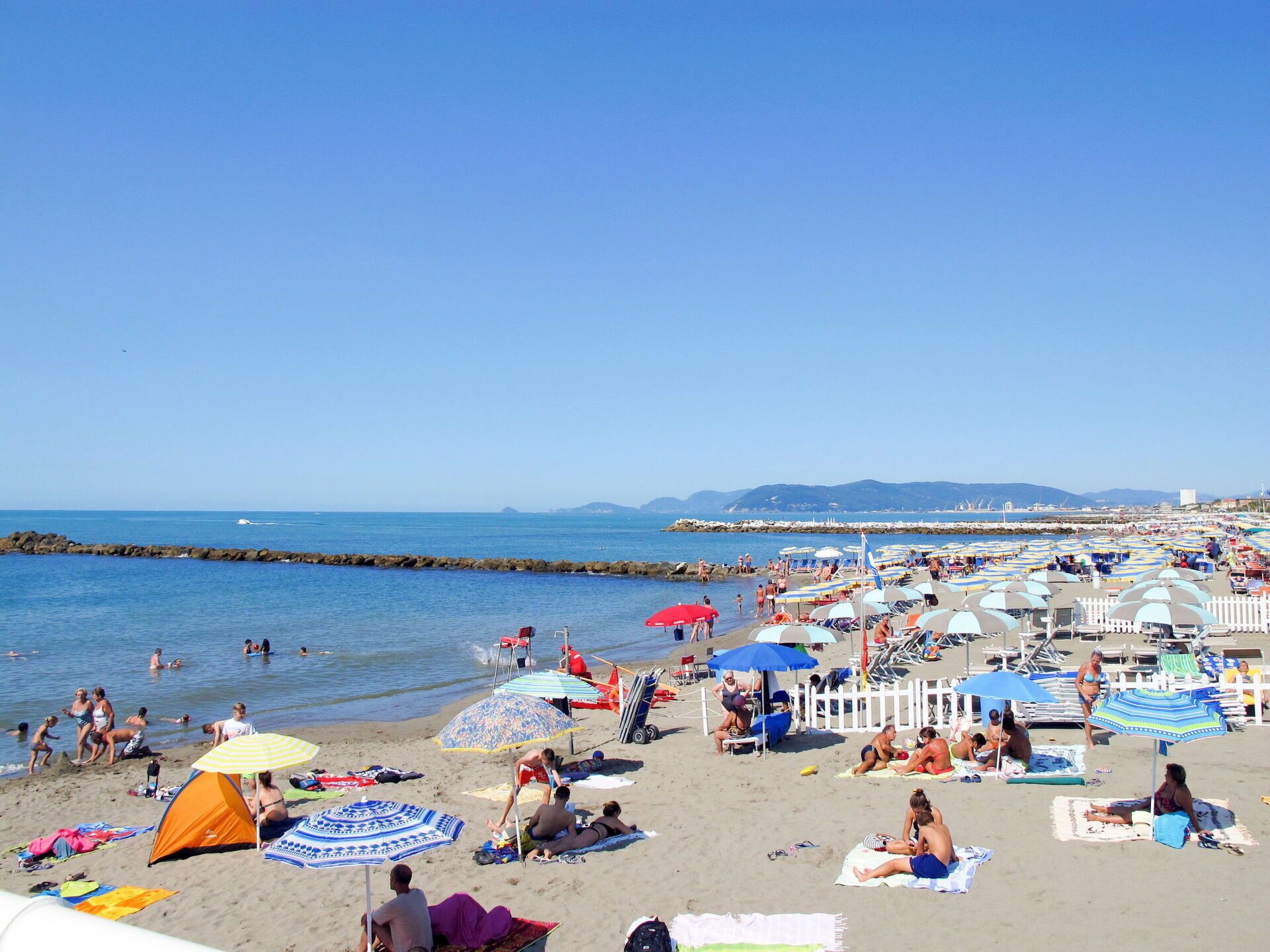 Una playa con sombrillas, gente tomando el sol y un rompeolas rocoso al fondo.