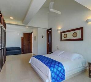 Ein Hotelzimmer mit Bett, Deckenventilator, blau gemustertem Bettbezug, dunkelblauem Sofa und einer Holztür.