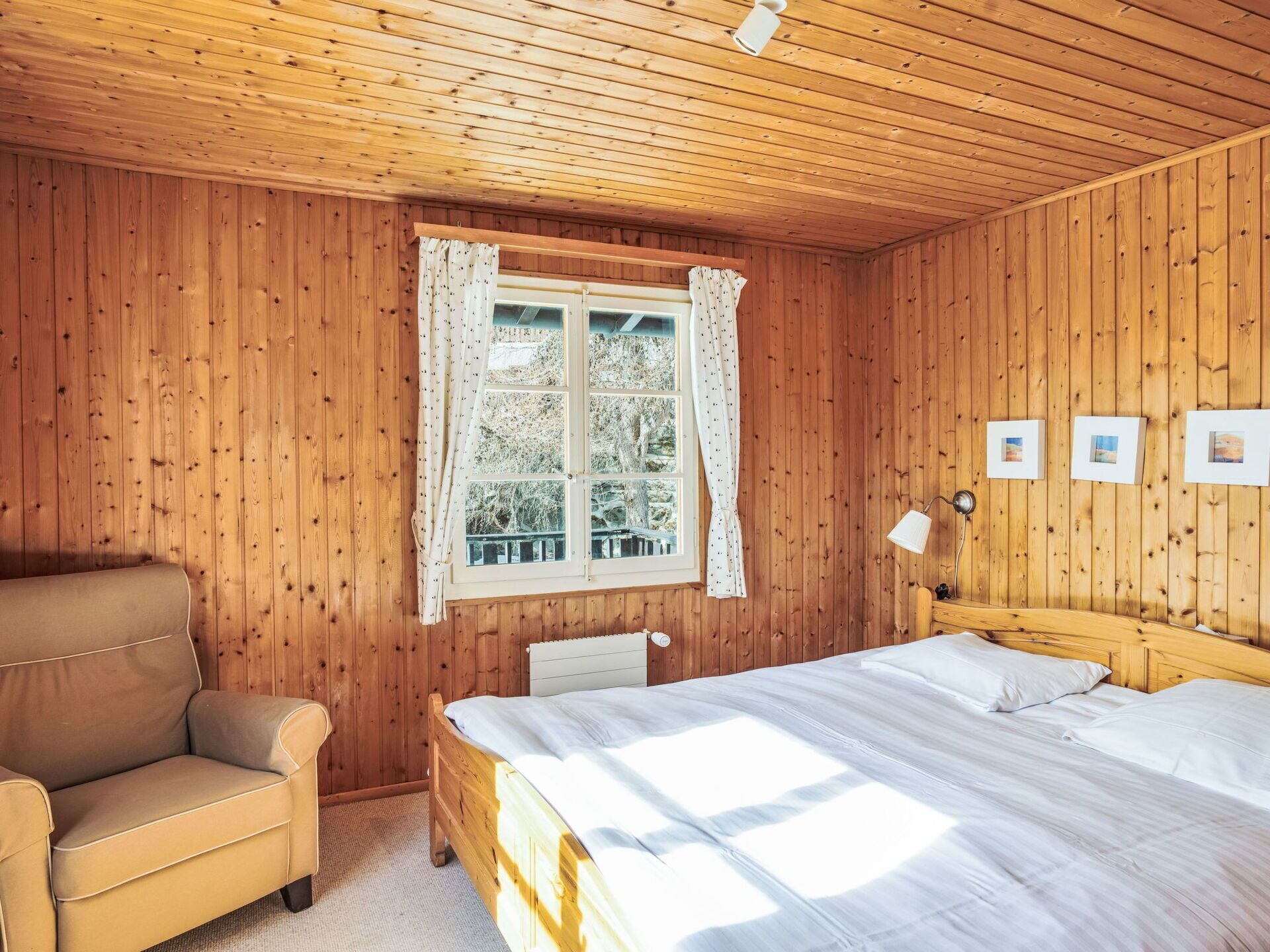 A wooden-paneled room with a bed, a chair, a window with curtains, and framed pictures on the wall.