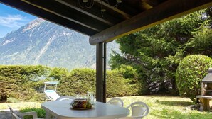 Area pranzo all'aperto con tavolo imbandito per due, circondata da sedie, con vista su un paesaggio montuoso.