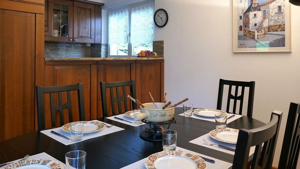 Zona pranzo con tavolo in legno apparecchiata per quattro persone, circondata da sedie, con una cucina sullo sfondo.
