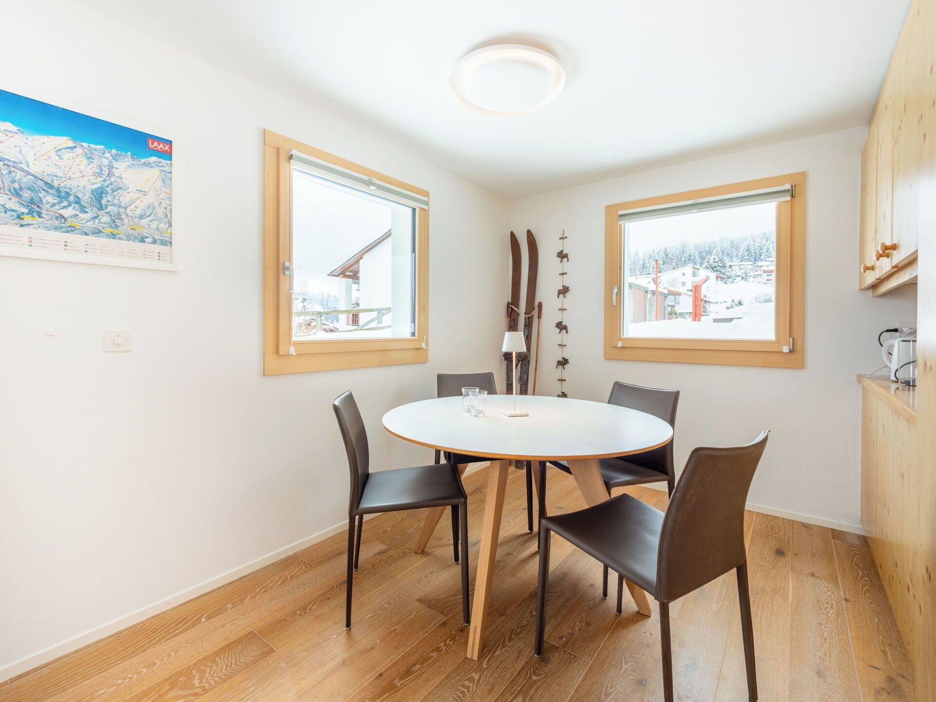 A dining area with a round table and chairs, two windows with wooden frames, a ceiling light, and a ski map on the wall.