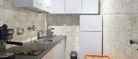 Una cocina compacta con armarios blancos, un lavamanos de acero inoxidable, una cafetera negra, un refrigerador blanco y un taburete de madera.