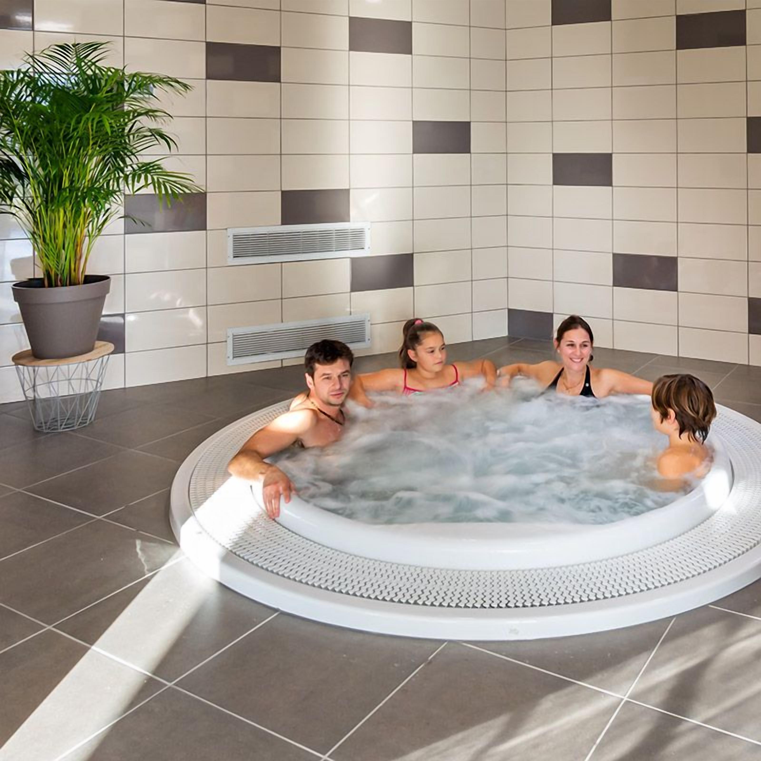 Un bain à remous où se trouvent quatre personnes, dans un décor intérieur moderne avec des murs carrelés et une plante en pot.
