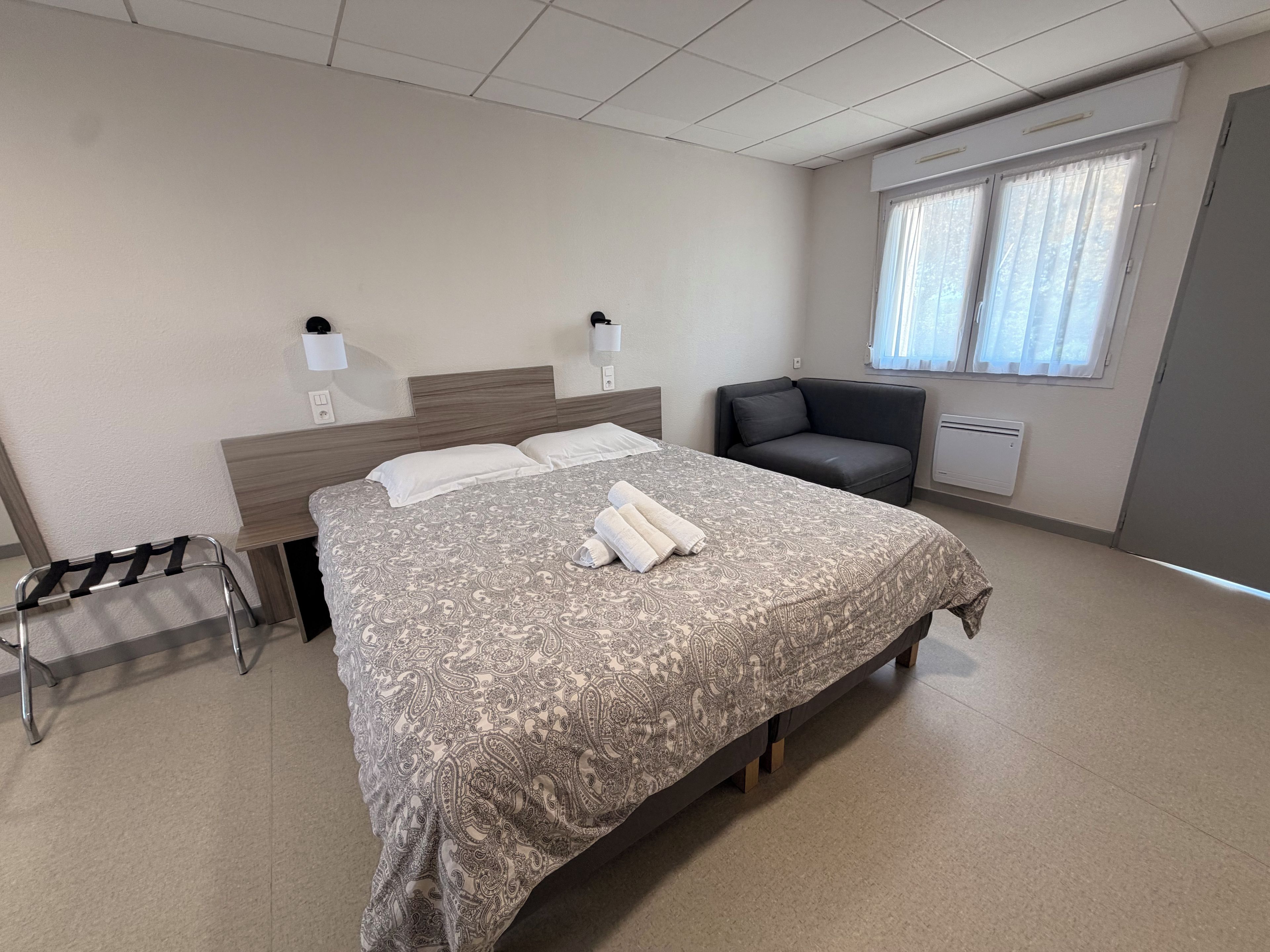 A hotel room with a bed, bedside lamps, a chair, a window with curtains, and a radiator.