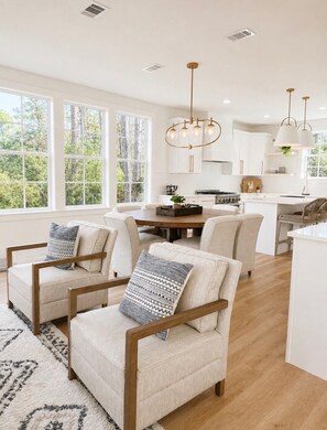 A modern kitchen with white cabinets, a dining table, and chairs. There are large windows providing natural light and a view of trees. - Escape to Paradise along the Emerald Coast (Rosemary Beach)