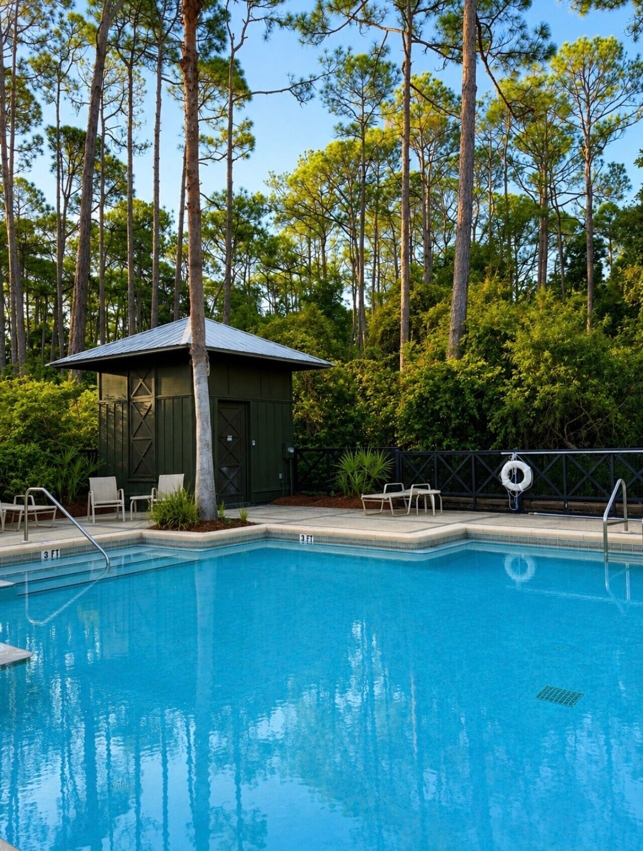 A rectangular swimming pool with a lifeguard stand and lounge chairs, surrounded by tall trees and a small building with a sloped roof.