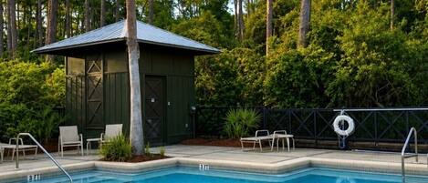 A rectangular swimming pool with a lifeguard stand and lounge chairs, surrounded by tall trees and a small building with a sloped roof.