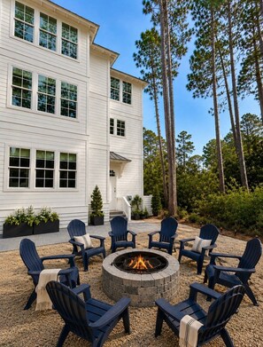 Une maison blanche à deux étages avec un foyer extérieur entouré de fauteuils Adirondack.
