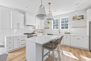 Une cuisine moderne avec des meubles blancs, un îlot central, des tabourets de bar, une cuisinière et des luminaires suspendus.