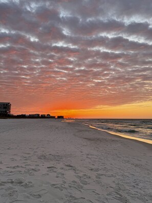Ein Strand mit Fußspuren, die zum Wasser führen, eine entfernte Küstenlinie mit Gebäuden und ein Sonnenuntergangshimmel.