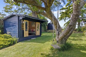A small wooden cabin with an open door, a swing, and a garden area. - Beautiful Cottage near Goodwood Estate (East Lavant)