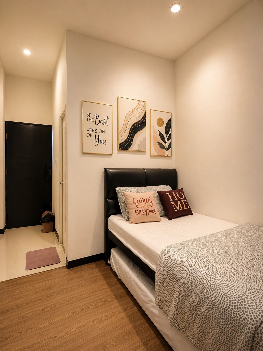 Un dormitorio con una cama, una pared con tres obras de arte enmarcadas, una puerta negra y un cesto de basura pequeño.