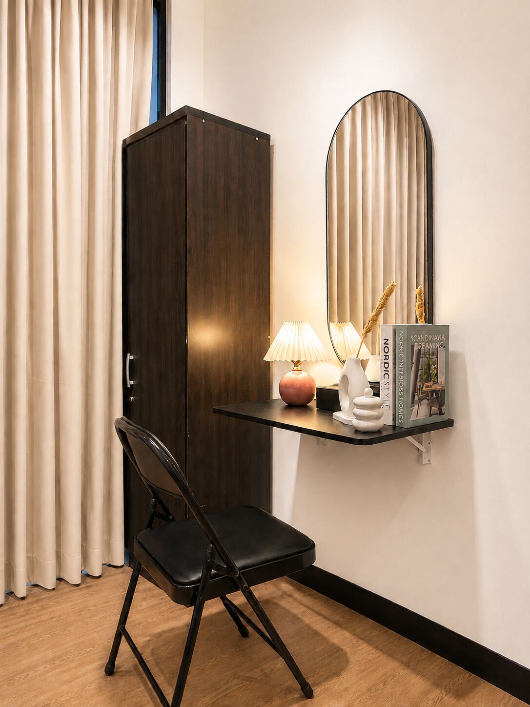 A room with a dark wooden wardrobe, a black chair, a small table with a lamp, a decorative vase, and a framed book on a shelf.