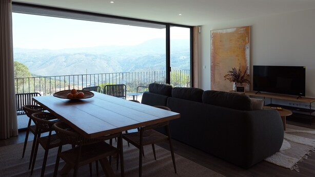 A modern living room with a large window, a dining table, and a sofa. - Casa Roca do Touro: Mountain View, Wi-Fi & Air Conditioning (Cabril)