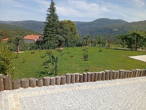 A grassy area with a wooden fence, a paved walkway, and a view of distant hills. - Casa Roca do Touro: Mountain View, Wi-Fi & Air Conditioning (Cabril)
