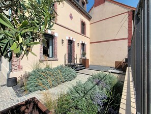 A courtyard with a brick pathway, a small garden with various plants, and a building with a beige facade and red brick accents. - La Maison du Port – Beach within walking distance, peaceful and comfortable (Dives-sur-Mer)