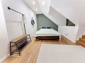 A modern bedroom with a sloped ceiling, a bed with a white bedspread, a bench, a mirror, and a staircase. - La Maison du Port – Beach within walking distance, peaceful and comfortable (Dives-sur-Mer)