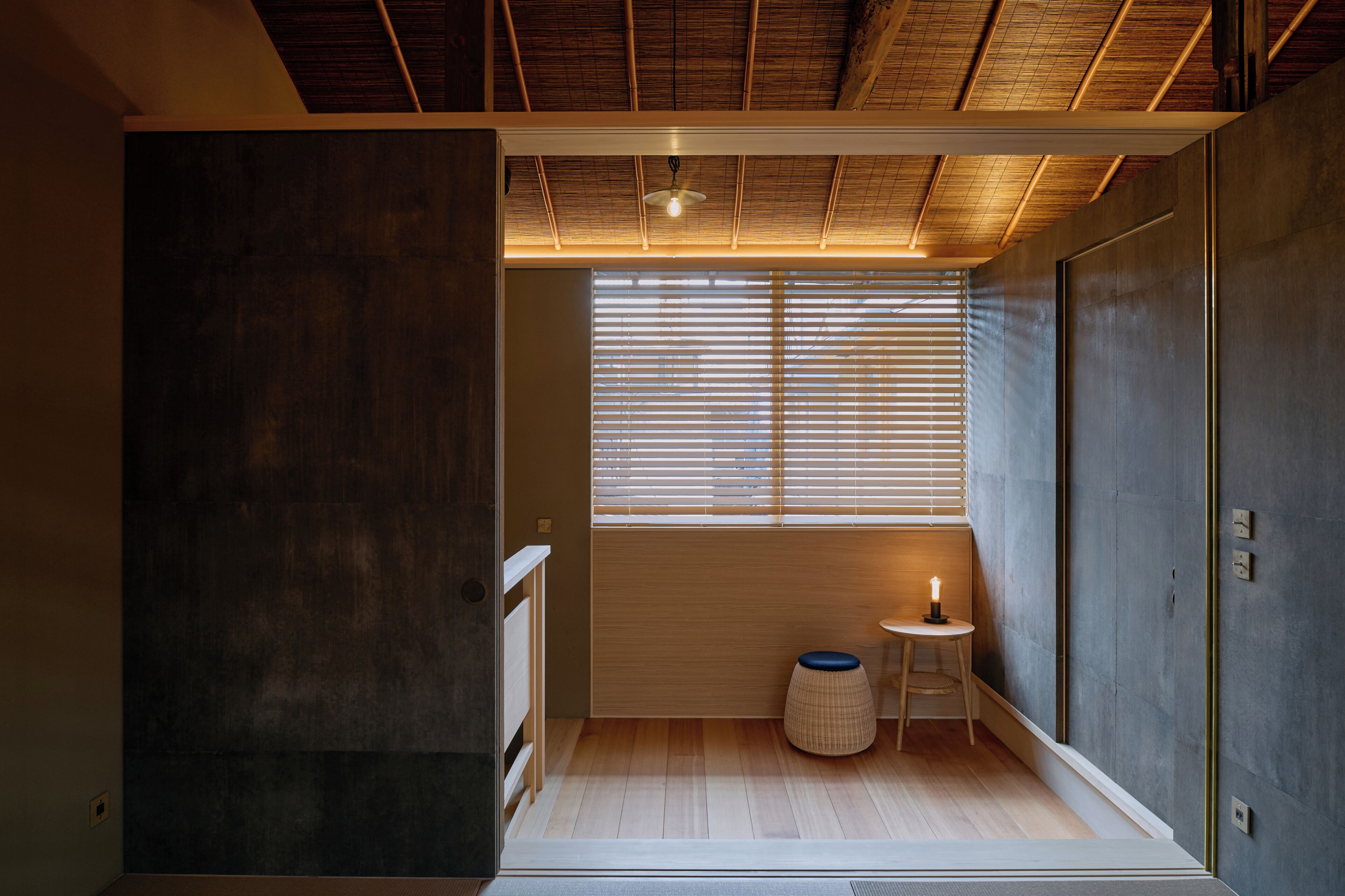 Een kamer met een houten vloer, een raam met jaloezieën, een kleine ronde tafel met een kaars, en een muur met een lichtschakelaar.