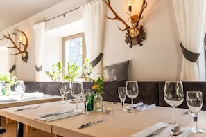 Een eetgedeelte met een lange tafel gedekt voor een maaltijd, decoratieve opbergplaats voor gewei aan de muur, en een raam met gordijnen.
