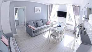 A modern living room with a grey sofa, a dining table with chairs, and a kitchen area with white cabinets.