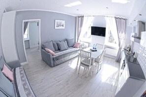 A modern living room with a grey sofa, a dining table with chairs, and a kitchen area with white cabinets.