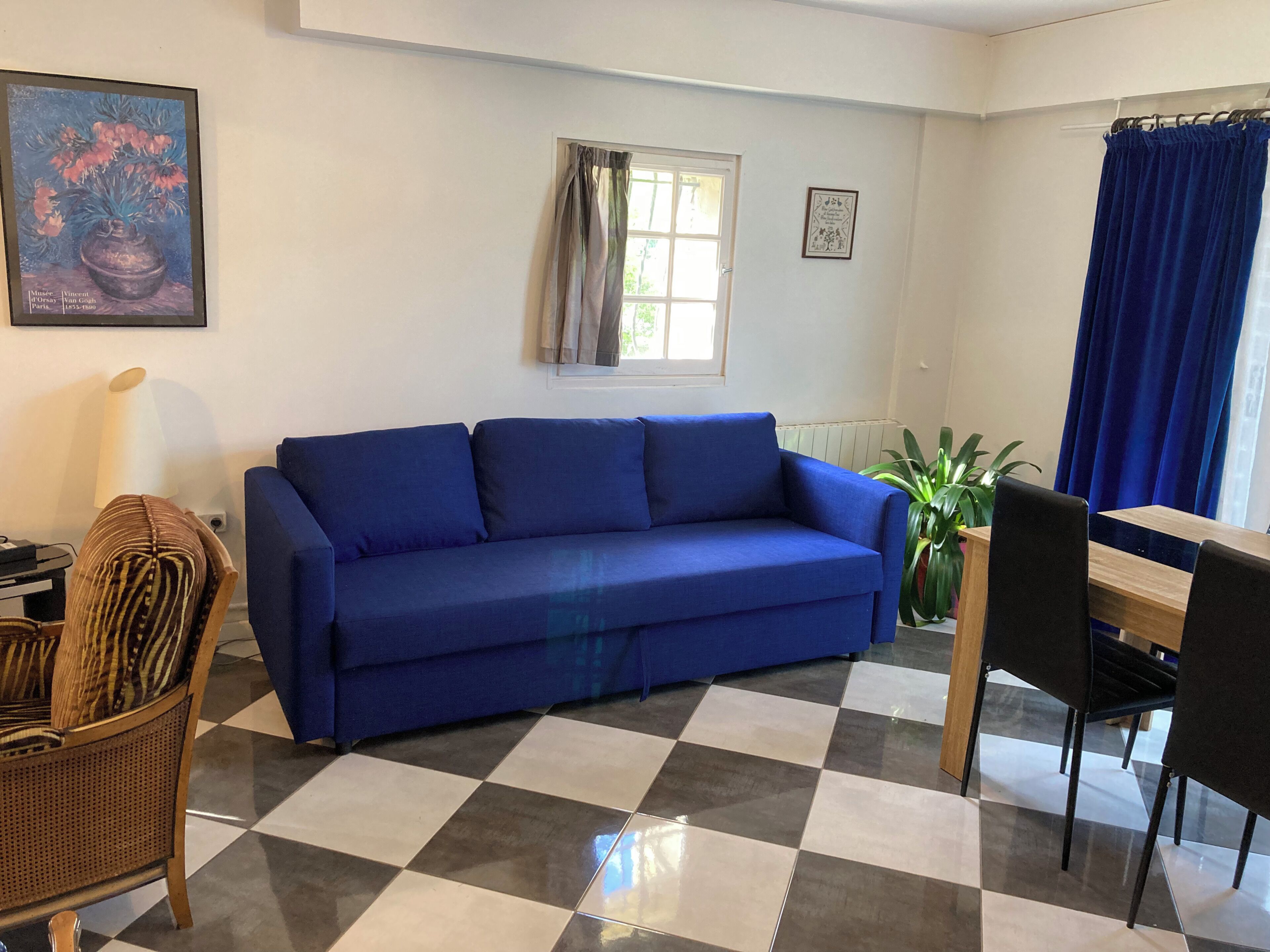 Una sala de estar con un sofá azul, una mesa de comedor con sillas, piso a cuadros, una ventana con cortinas y un cuadro enmarcado en la pared.