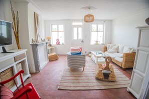 A living room with a television, wicker furniture, a striped ottoman, and a red chair. - La Parenthèse d’Honfleur Cosy | 2 Bedrooms & Private Courtyard (La Rivière-Saint-Sauveur)