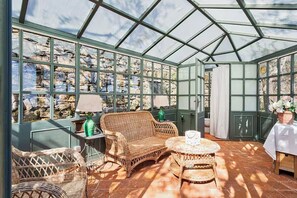 Ein Wintergarten mit Korbmöbeln, einem runden Tisch und Blick auf eine Steinmauer.