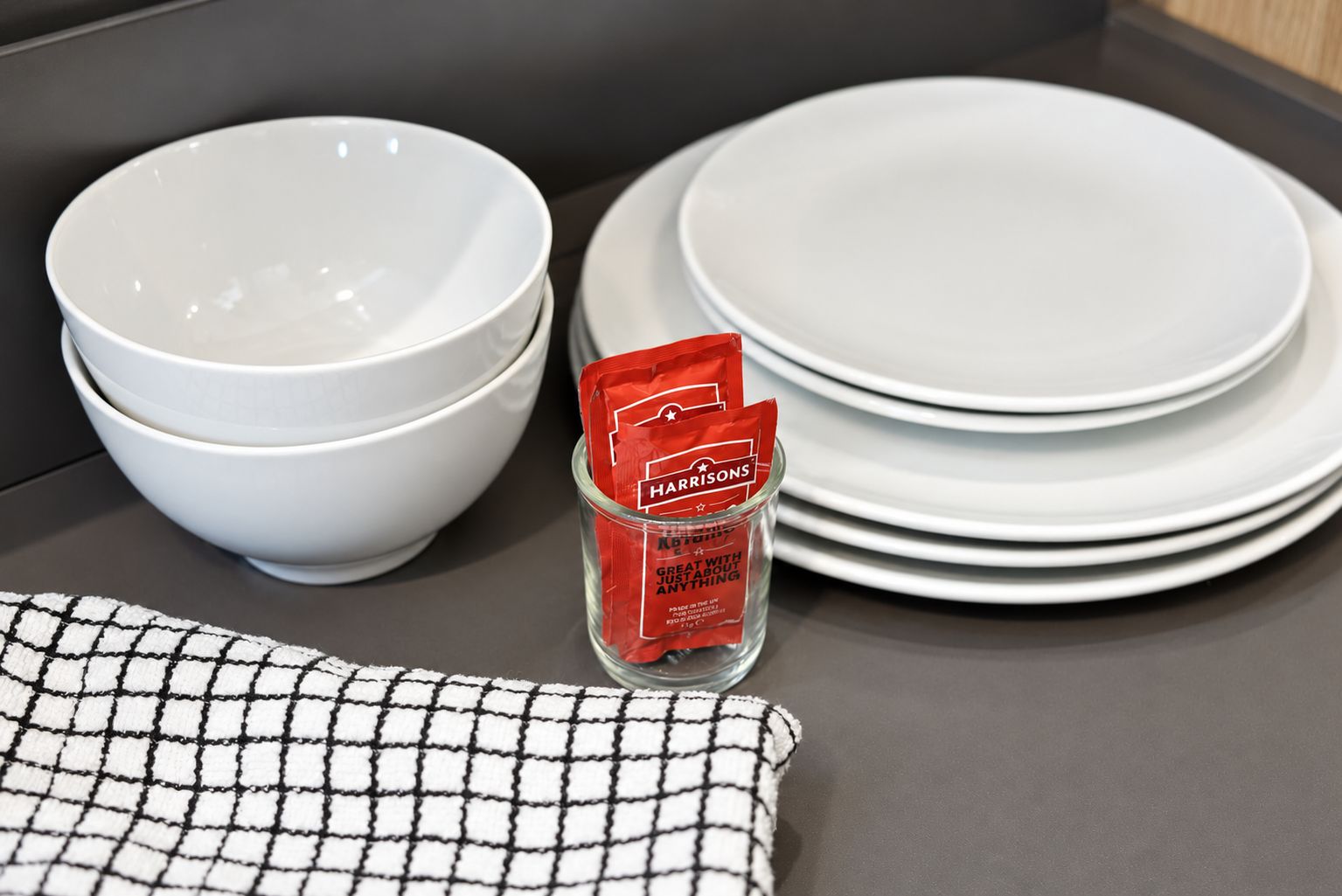 White bowls stacked on a table with a glass jar containing a red packet labeled 'HARRISONS' and a black and white checkered cloth.