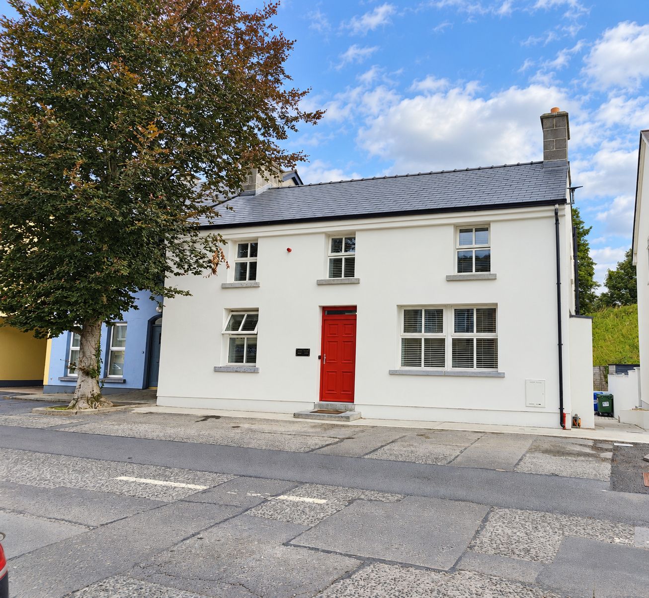 Un bâtiment blanc à deux étages, avec une porte rouge, de nombreuses fenêtres et un arbre devant.