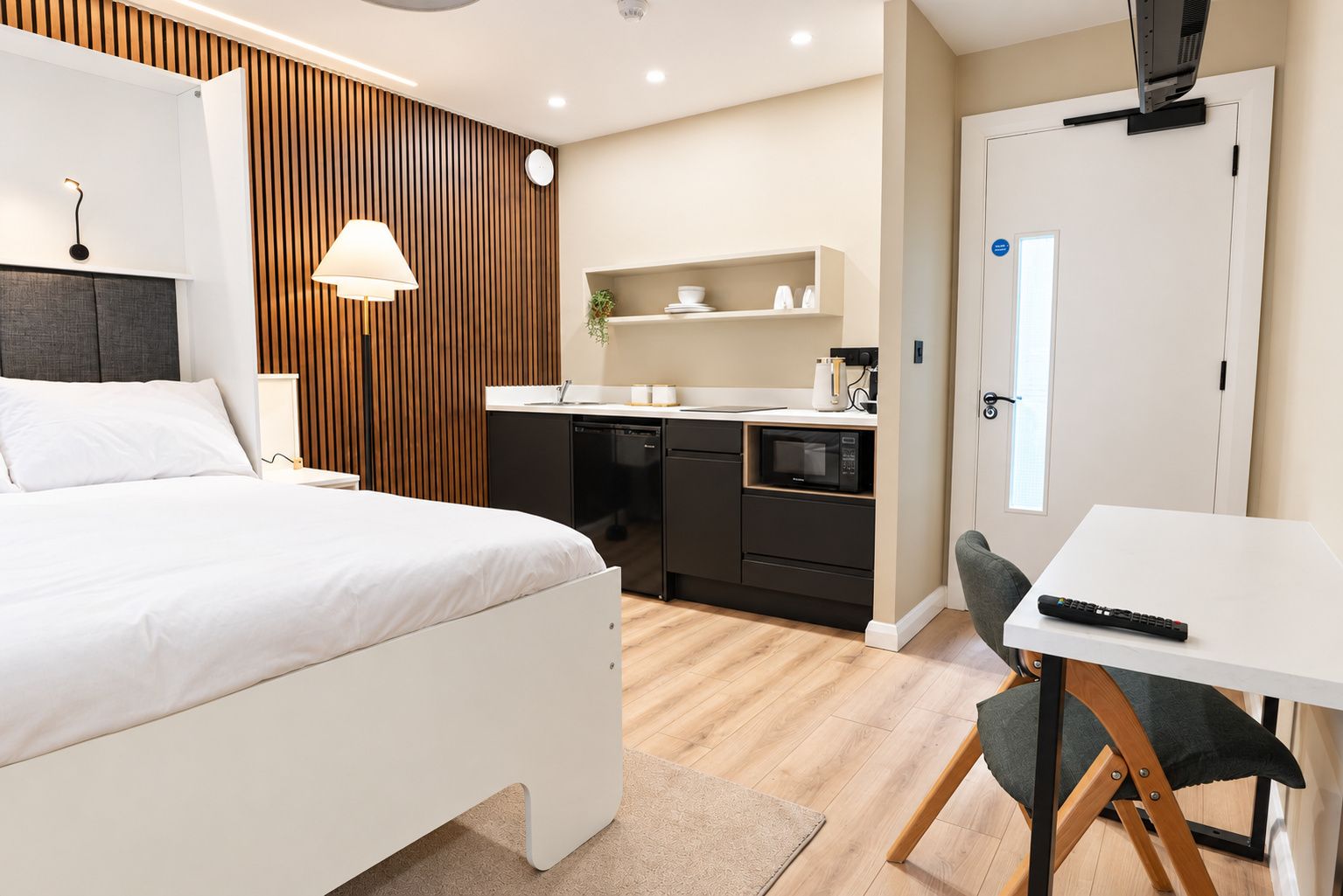 A modern hotel room with a bed, bedside table, lamp, kitchenette, and a small desk with a chair.
