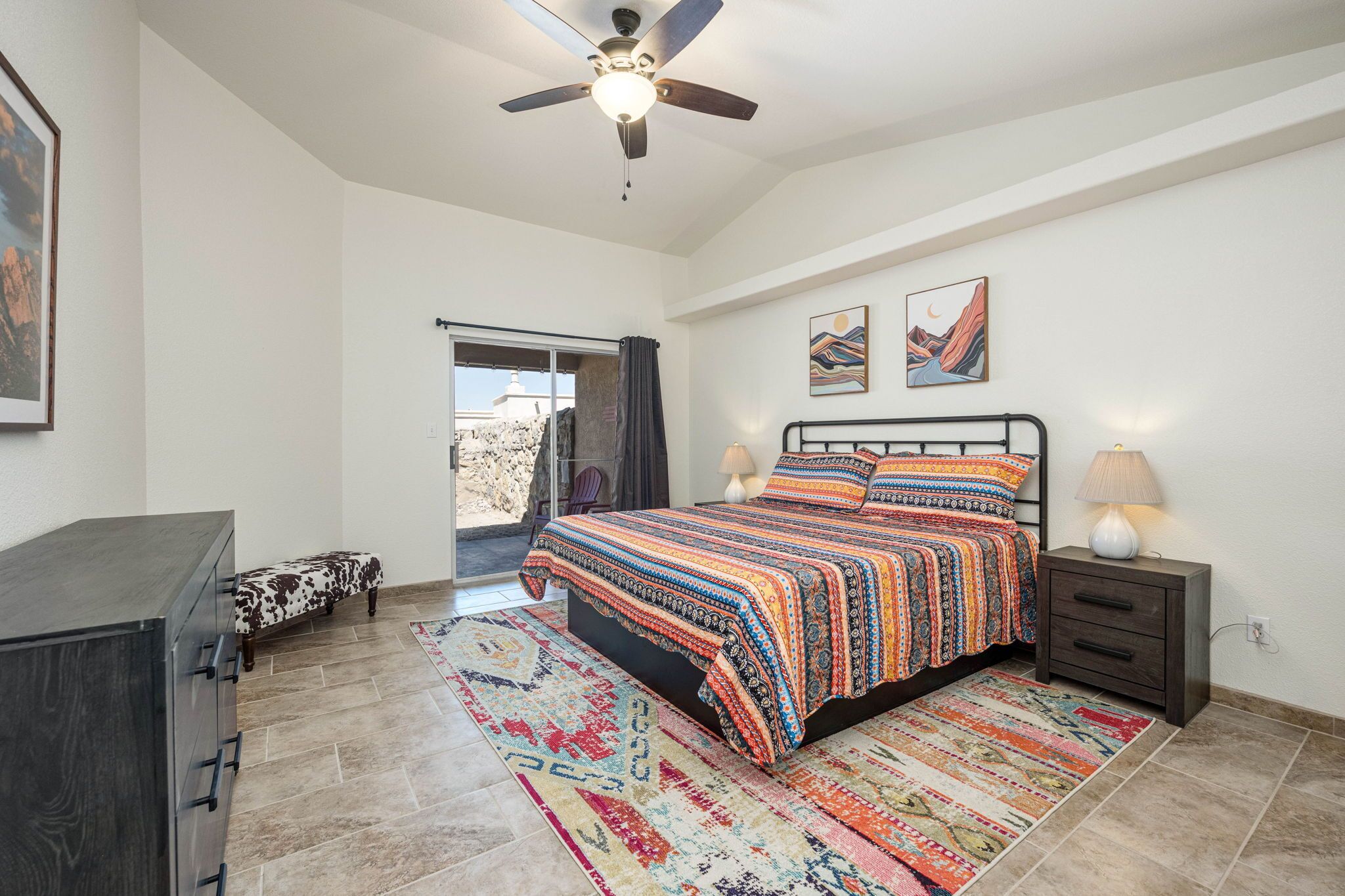 Une chambre à coucher comprenant un lit, une table de chevet, une commode, un ventilateur de plafond et une vue sur l’extérieur, visible à travers une porte vitrée.