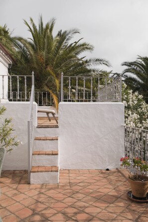 Un espace extérieur aux murs blancs, doté d’un escalier menant à un niveau supérieur, et entouré d’une clôture en métal et de palmiers.