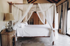 Una cama con dosel y cortinas, ropa de cama blanca, una mesita de noche de madera con una lámpara y un tapete tejido sobre suelo de baldosas.