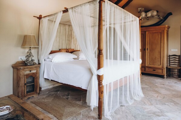 Un lit à baldaquin avec du linge de lit blanc, une table de chevet en bois avec une lampe et un sol en pierre.