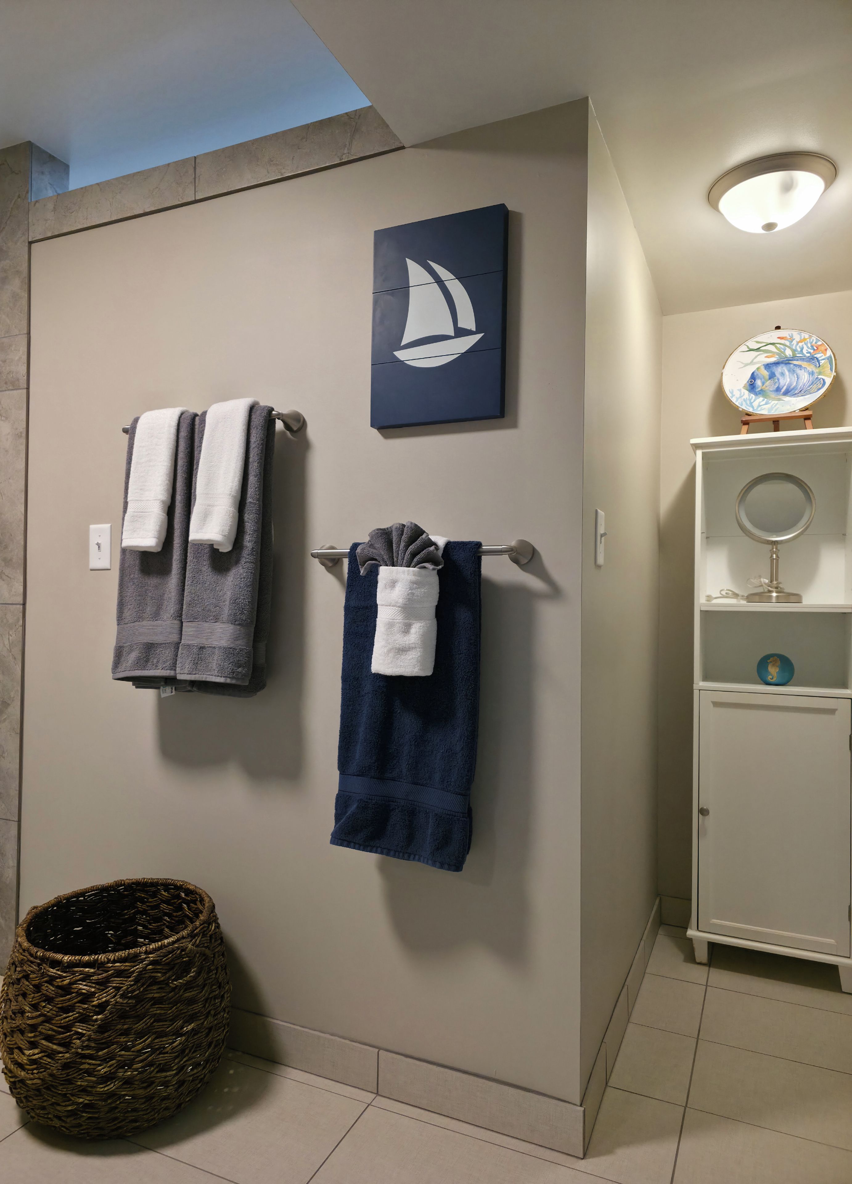 A bathroom with two towels hanging on a towel rack, a woven basket on the floor, a wall-mounted shelf with a decorative plate, and a ceiling light fixture.
