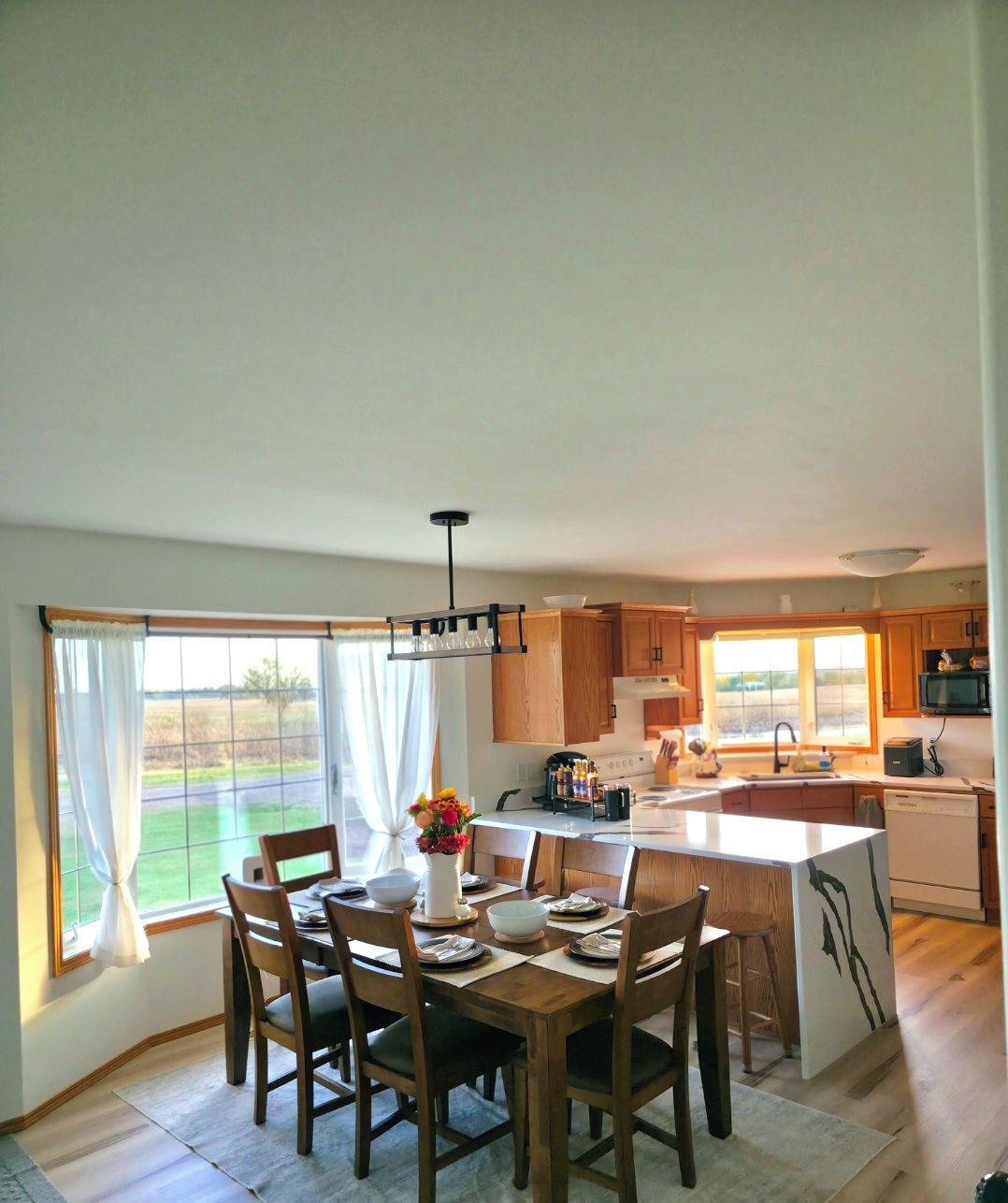 A modern kitchen with wooden cabinets, a dining table set for four, and a large window providing a view of the outdoors.