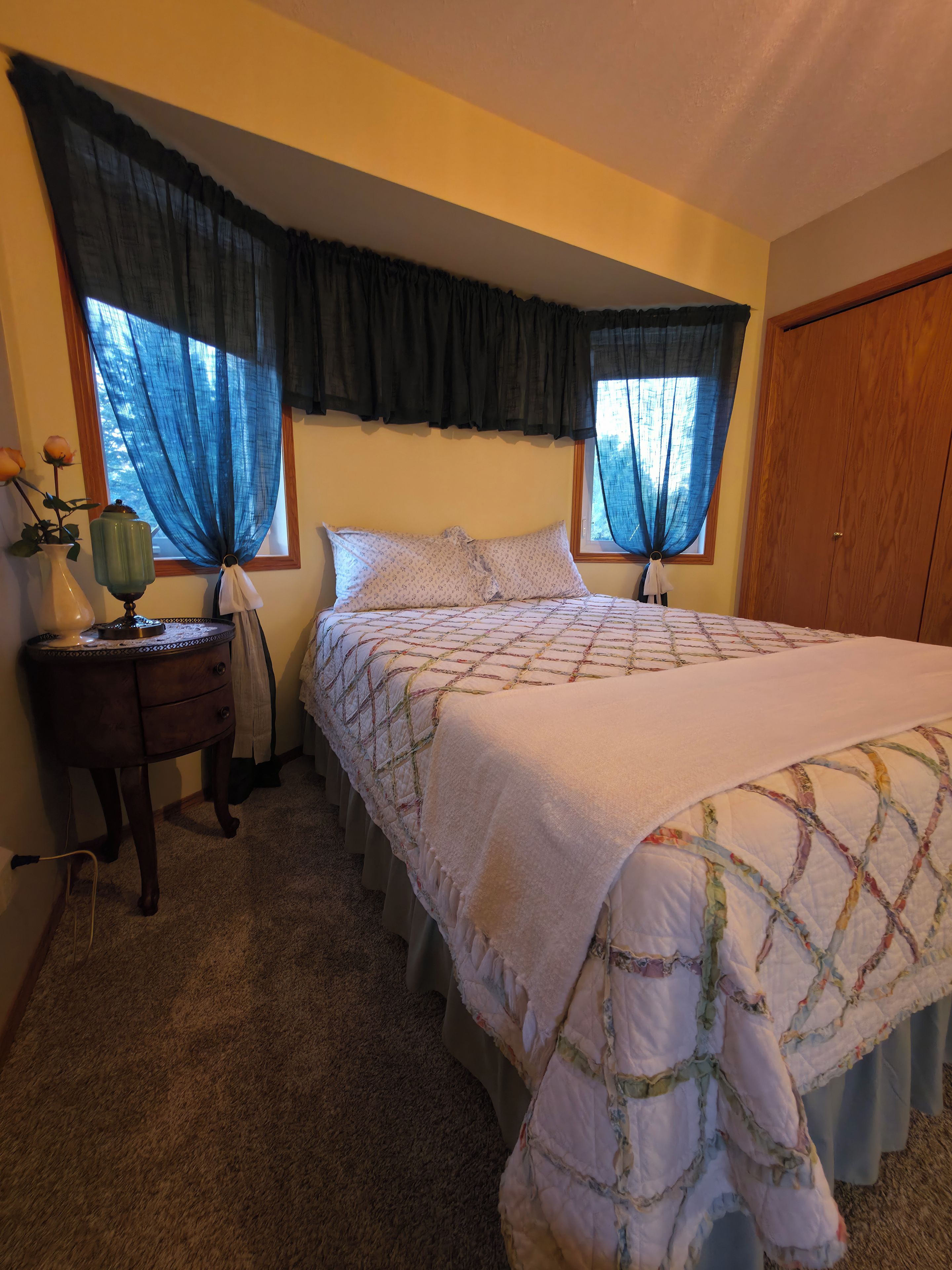 Une chambre à coucher avec un grand lit, deux fenêtres avec des rideaux, une table de chevet avec une lampe et une armoire en bois.
