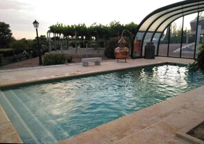 A swimming pool with a curved glass enclosure, surrounded by a landscaped garden with a lamp post and a large copper pot.