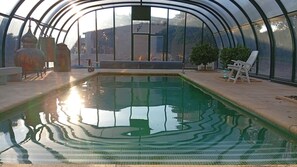 A swimming pool with a curved glass enclosure, surrounded by outdoor furniture and potted plants.