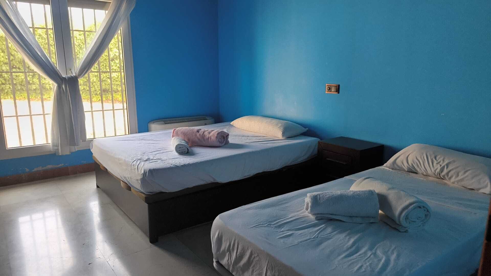 Una habitación con dos camas, una ventana con cortinas, una mesita de noche y un interruptor de luz en la pared.