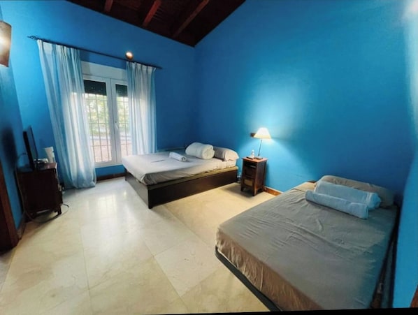 A room with two beds, a window with curtains, a bedside table with a lamp, and a small wooden cabinet.