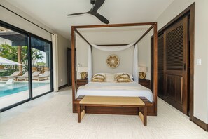 Uma cama com dossel e cortina, duas mesas de cabeceira, um objeto decorativo na parede e vista para uma área de piscina externa.