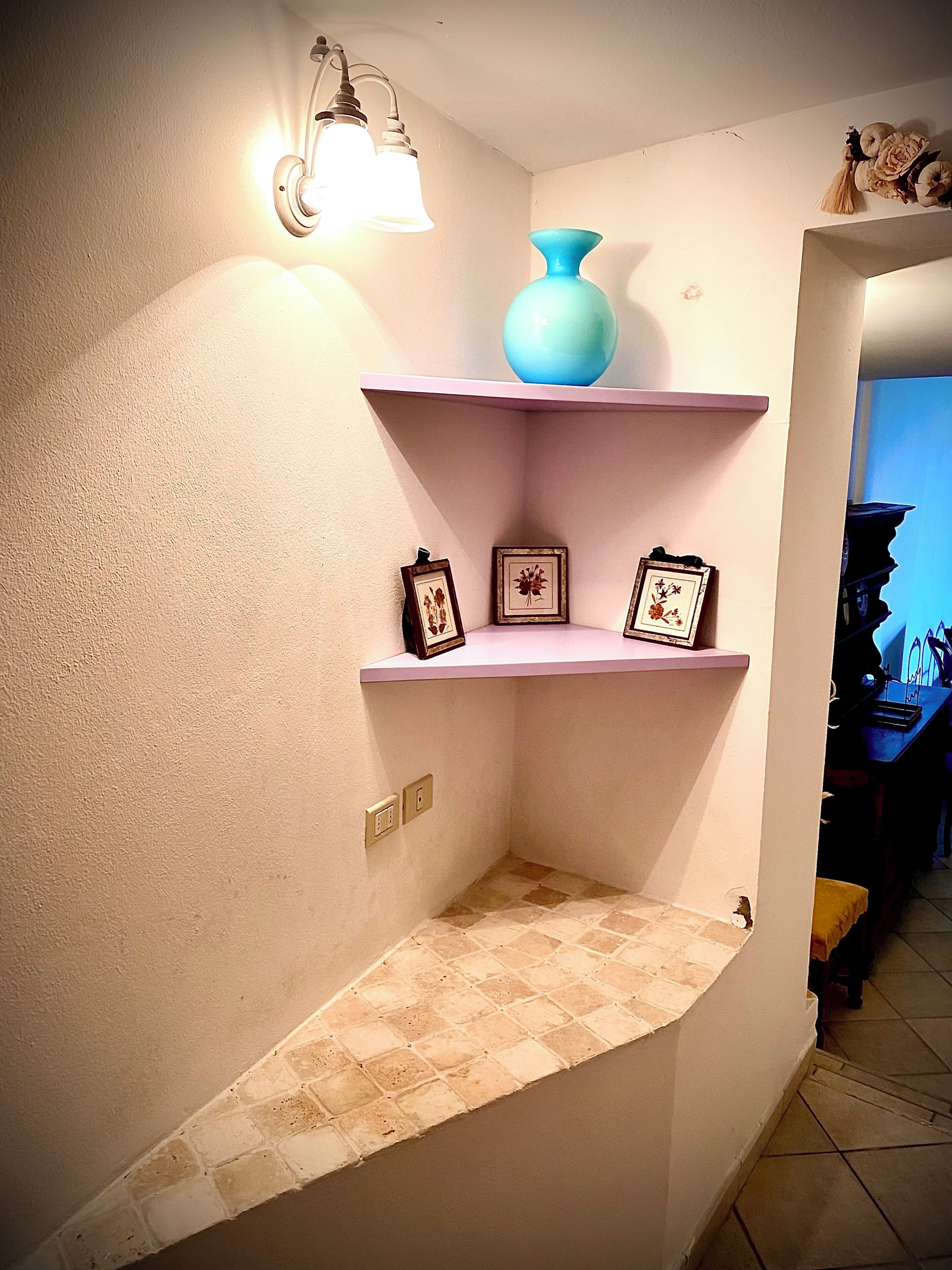 En hörna med en lampa, en blå vas och inramade bilder på hyllor.